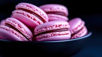 Pink French macarons with cream filling arranged in black ceramic bowl against dark background, close up view showing delicate meringue texture and filling.