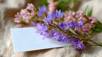 Purple and pink wildflowers lying on blank white paper card against natural linen textile background, soft morning light creates gentle shadows.
