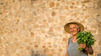 Senior woman with light skin tone, wearing straw hat and apron, holding fresh green vegetables against rustic stone wall background for gardening and farming concepts.