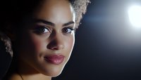 Young African American woman with natural curly hair smiling confidently in dramatic side lighting, wearing burgundy lipstick against dark background with lens flare effect.