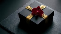 Elegant black gift box with golden ribbon and red orchid flower on dark slate surface for luxury present wrapping and special occasions.