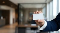 Businessman in dark suit holding blank white business card in modern office corridor with glass partitions and natural light from windows.