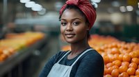 Young African American woman in apron and red headscarf smiling while working at produce section with fresh oranges in grocery store background, retail employee portrait.