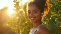 Young African American woman with natural curly hair smiling outdoors at golden sunset, wearing gray tank top against green foliage background with sun flare effect.