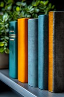 Row of hardcover books in teal blue, orange and gray colors standing on wooden shelf with green plant in background, creating modern home library atmosphere.