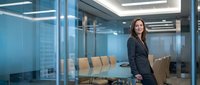 Professional businesswoman in dark suit standing confidently in modern glass conference room with city skyline view for corporate presentations.