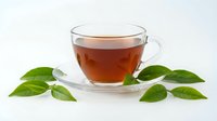Clear glass teacup with amber tea on white background surrounded by fresh green leaves for beverage and wellness designs.