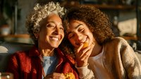 Joyful senior woman with gray curly hair and young adult female with natural curls sharing intimate moment in warm sunlight.