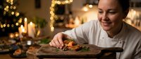 Professional chef plating gourmet dish in warm restaurant kitchen with festive lights creating cozy dining atmosphere for holiday celebration.