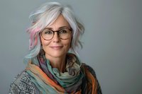 Mature woman with stylish white-pink hair, glasses and colorful scarf smiling warmly against gray background. Natural beauty and confidence in aging gracefully.