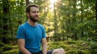Caucasian male in blue t-shirt sitting on moss-covered ground in sunlit forest, contemplative expression, outdoor nature setting for wellness and lifestyle content.