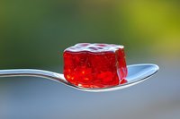Red gelatin dessert cube on silver spoon against blurred green background, showcasing translucent texture and vibrant color for food photography.