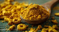 Ground turmeric powder in wooden spoon with raw turmeric root pieces scattered on rustic green surface, macro food photography in warm lighting.