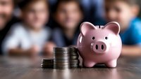 Pink ceramic piggy bank next to stacked silver coins on wooden surface, symbolizing savings and financial education with blurred students in background.