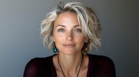 Young woman with platinum blonde wavy hair and turquoise earrings wearing black mesh top against gray background, natural makeup and confident expression.