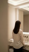 Young Asian woman with long dark hair in white shirt looking at bathroom mirror in modern luxury hotel suite with marble countertop and warm lighting.