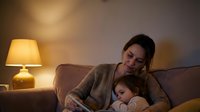 Young mother in cozy sweater bonding with sleeping toddler daughter on living room sofa in warm evening light for family lifestyle content.