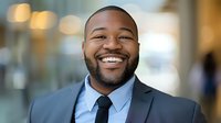 Confident African American businessman with genuine smile wearing suit and tie in corporate office setting with blurred background and natural lighting.