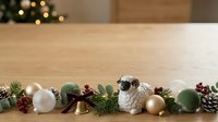 Festive holiday decorations arranged in a row on wooden surface, featuring white and green ornaments, gold bells, evergreen branches, pinecones, and ceramic sheep figurine for seasonal home decor.