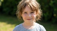 Young blonde girl with curly hair smiling outdoors in striped shirt on sunny day, childhood happiness and carefree summer moments in natural setting.