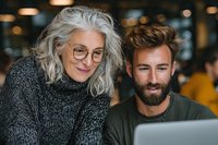 Senior woman with glasses and gray hair in sweater smiling while looking at laptop with bearded young man in casual setting, representing intergenerational collaboration.