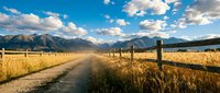 Rural dirt road through golden wheat field with wooden fence leading to majestic mountain range under dramatic cloudy blue sky in countryside landscape.