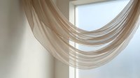 Sheer beige curtain fabric draped elegantly across a bright window, creating soft flowing layers with natural light filtering through for interior design and home decor projects.