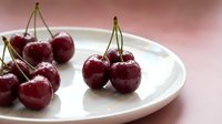 Fresh ripe red cherries with stems arranged on white ceramic plate against soft pink background for healthy eating and summer fruit concepts.