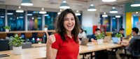 Happy businesswoman giving thumbs up in modern coworking office space with colleagues working in background during daytime.