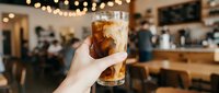 Caucasian hand holding glass of cola with ice in casual cafe setting with warm string lights and blurred interior background, suitable for beverage, lifestyle, and hospitality marketing.
