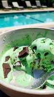 Mint chocolate chip ice cream in bowl with spoon by poolside, lounge chairs and turquoise water visible in background for summer dessert and lifestyle content.