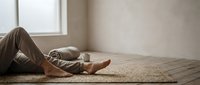 Young woman relaxing on woven rug in sunlit room with bare feet and casual clothing for wellness and mindfulness concepts.