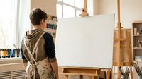Young male artist in apron standing at easel with blank canvas in bright art studio workspace ready for creative painting session.
