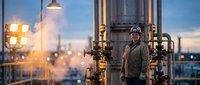 Middle-aged Caucasian male industrial worker in safety helmet standing confidently at oil refinery facility during golden hour sunset with industrial equipment.