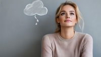 Young Caucasian woman in beige sweater with thoughtful expression and illustrated cloud thought bubble on gray wall background, representing contemplation and mindfulness.