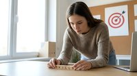 Young woman with dark hair and light skin tone, wearing a beige ribbed sweater, writing the word "skills" on white paper at wooden desk in bright office space with laptop and target board visible, representing professional development and career planning.