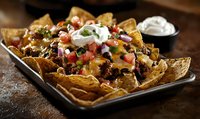 Loaded nachos topped with seasoned ground beef, melted cheese, fresh tomatoes, black beans, sour cream and garnished with chopped herbs on dark rustic platter.