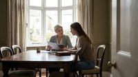 Two women collaborating at wooden dining table reviewing documents in bright home interior with large windows and natural lighting for business meeting.