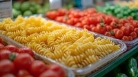 Fresh raw fusilli pasta in grocery store display with vibrant cherry tomatoes and green vegetables, selective focus on yellow pasta spirals in plastic container.