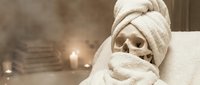 Skeleton wrapped in white gauze bandages with spa setting, candles and towels in soft-focus background for Halloween, medical education or wellness themes.