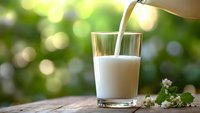Fresh milk being poured into glass on wooden table with green bokeh background and small white flowers for healthy breakfast concept.