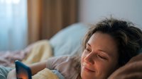 Woman with dark hair, appearing to be in her 40s of mixed ethnicity, relaxing on a bed with pillows and blankets in a cozy bedroom setting. Suitable for lifestyle, wellness, and home comfort content.