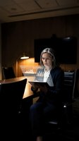 Middle-aged woman with long gray hair, Caucasian ethnicity, wearing dark business suit and white shirt, working late at office desk with laptop displaying financial charts and graphs, warm desk lamp illuminating workspace in dark modern office environment.