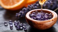 Pouring essential oil onto purple amethyst crystals in wooden bowl with orange and lavender, creating spa wellness atmosphere on dark background.