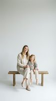 Young Caucasian mother and daughter sitting together on wooden bench wearing matching beige outfits against neutral background for family lifestyle content.