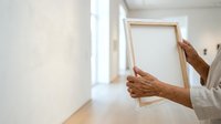 Senior man with brown skin tone holding wooden frame in bright hallway, wearing white shirt. Suitable for home renovation, interior design, and lifestyle content.