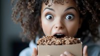 Young African American woman with curly hair expressing surprise while holding box of chocolate cookies, showing excitement and joy in candid moment.