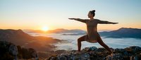 Young athletic woman practicing warrior yoga pose on mountain peak during golden sunrise with misty valleys below, outdoor fitness and mindfulness concept.