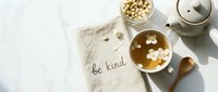 Jasmine tea in a white cup with floating flowers, be kind linen napkin, teapot, and wooden spoon on marble surface, top view.