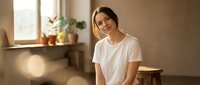 Young woman in white t-shirt sitting relaxed in bright modern kitchen with natural sunlight streaming through window creating warm atmosphere.